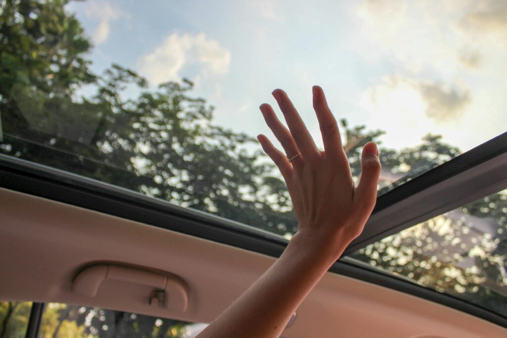 A hand reaching out through a car sunroof, touching the sky on a sunny day.