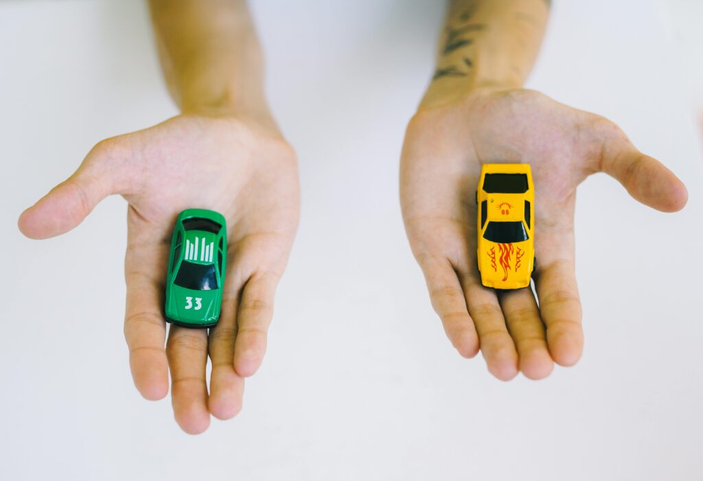 Pair of adult hands holding green and yellow toy cars on a white surface, symbolizing choice.